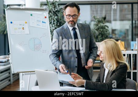 Focused business partners working on corporate project, discussing new plan. Stylish blonde-hair woman ask for advice about some documents or financial graphs from confidenr male colleague Stock Photo