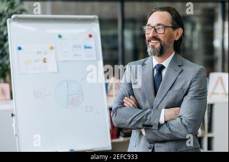 Influential serious ceo stands near marker board at the workplace, looks away with cross hands. Confident bearded middle aged business man wearing formal suit and eyeglasses Stock Photo
