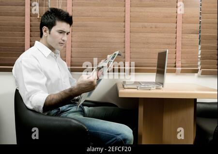 Side profile of a young man sitting in front of a laptop reading a newspaper Stock Photo