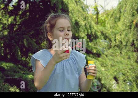 Portrait of a cute blonde young kid, girl blowing bubbles and smiling, with green trees background in the park. Copy space, front view, bokeh Stock Photo