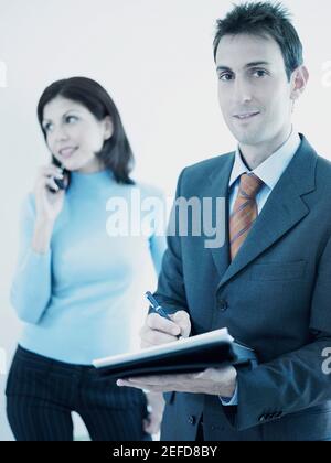 Portrait of a businessman writing on a personal organizer with a businesswoman standing behind him holding a mobile phone Stock Photo