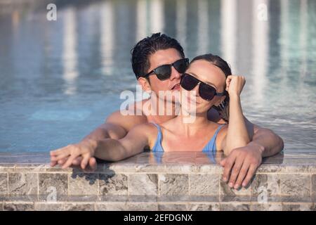 Very happy couple wearing sunglasses enjoy each other in swimming pool. Man holding woman on hands Stock Photo