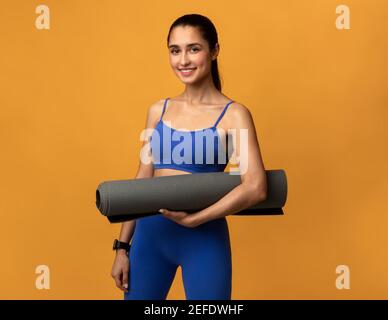 Wellness. Smiling millennial female athlete holding yoga mat standing isolated on orange studio background. Positive young woman promoting healthy lif Stock Photo