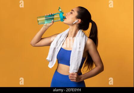 Fitness And Healthy Lifestyle Concept. Portrait of sporty young woman drinking mineral water from bottle at studio after workout at gym, wearing earbu Stock Photo