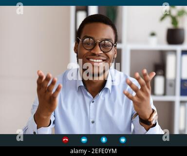 Cheerful african american guy in glasses talking and gesturing, office interior, copy space, pov screen. Positive black man speaker having online conf Stock Photo