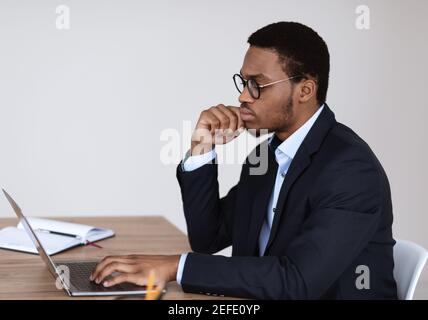 Pensive african american businessman in glasses working with laptop in office, looking at computer screen and touching his face, looking for creative Stock Photo