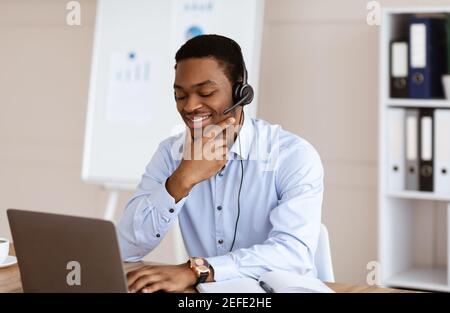 Positive young black man having online business conference in office, using laptop and headset, cheerfully talking to client or business partner, look Stock Photo