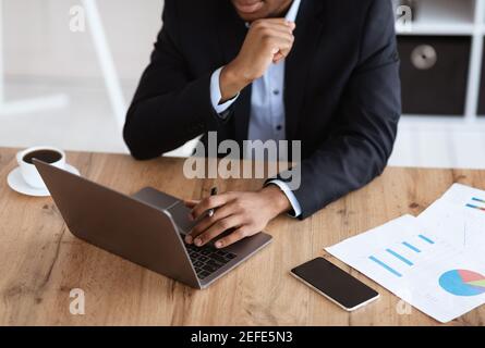 Cropped of black businessman in suit working with laptop in office, checking marketing report, modern technologies in business concept. Unrecognizable Stock Photo