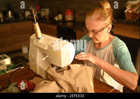 Confident senior elderly woman seamstress white shirt and glasses sitting in front of sewing machine, working on clothes at home using beige fabric. Retired people, age, job occupation concept Stock Photo