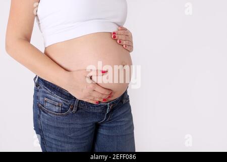 Pregnant woman in jeans and white top hold hands on her belly, casual minimalistic pregnancy Stock Photo