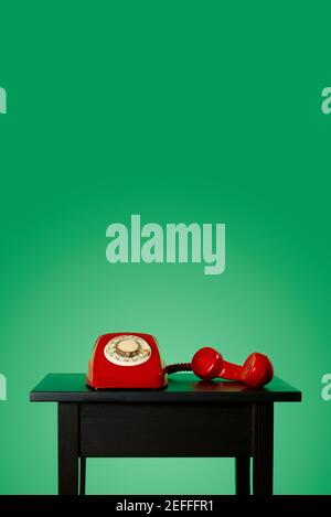 red landline rotary dial telephone, with its handset off the hook on a black wooden table, on a green background with some blank space on top Stock Photo