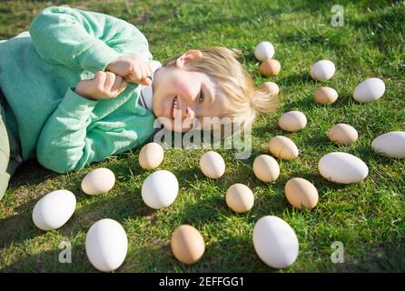 many white and beige chicken and goose eggs lie on the bright green grass around the head of a cheerful, happy 3-4 year old blond boy. On the farm, pr Stock Photo