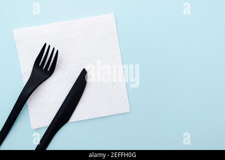 Set of plastic cutlery black fork and knife on white paper napkin and  blue background, top view, space for text. Stock Photo