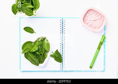 Composition with spinach, measuring tape, blank paper notebook and pink alarm clock on white background. Mock up. Diet concept, copy space