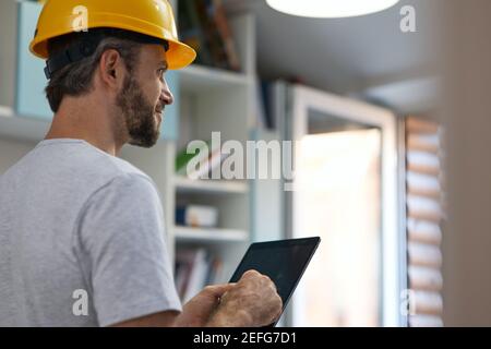 Professional repairman wearing helmet looking aside, using digital tablet while standing indoors Stock Photo