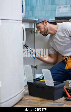 Professional repairman looking focused, using pipe wrench while examining and fixing sink pipe in the kitchen Stock Photo