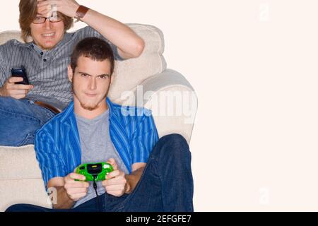 Young man playing video game with another young man using a mobile phone behind him Stock Photo