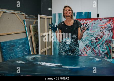 Young woman paints an abstract picture with her hands and spatula on a work table in her interior studio. Focus on hand Stock Photo