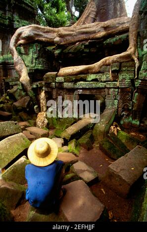 High angle view of a man crouching on a stone in front of a temple, Ta Prohm Temple, Angkor, Siem Reap, Cambodia Stock Photo