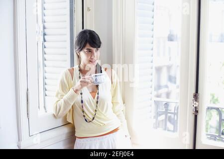 Young woman leaning against a window and holding a cup of tea Stock Photo