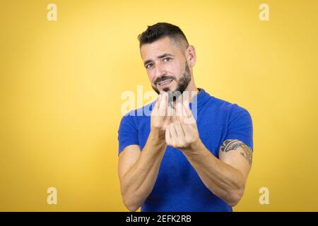Handsome man with beard wearing blue polo shirt over yellow background doing money gesture with hands, asking for salary payment, millionaire business Stock Photo
