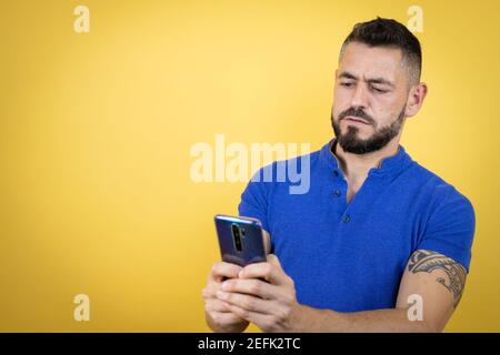 Handsome man with beard wearing blue polo shirt over yellow background chatting with his phone Stock Photo