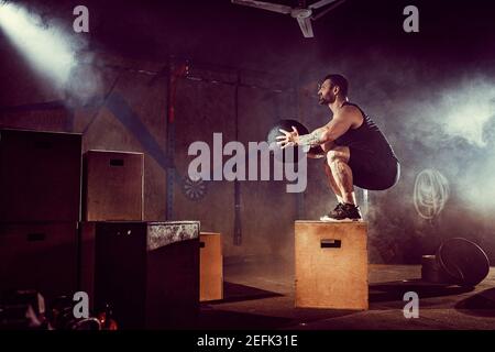 Athlete gave exercise. Jumping on the box. Phase touchdown. Gym shots in the dark tone. Smoke in gym. Stock Photo
