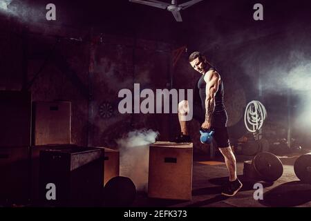 Muscular attractive caucasian bearded tasttoed man lifting kettlebell in a gym. Weight plates, dumbbell and tires in smoke background. Stock Photo