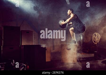 Athlete gave exercise. Jumping on the box. Phase touchdown. Gym shots in the dark tone. Smoke in gym. Stock Photo