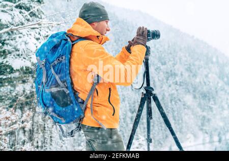 Photographer dressed orange softshell jacket with backpack making a landscape shoot using a digital camera and tripod while he trekking winter mountai Stock Photo