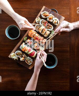 Overhead of hands using chopsticks to pick up sushi rolls off platter. Stock Photo