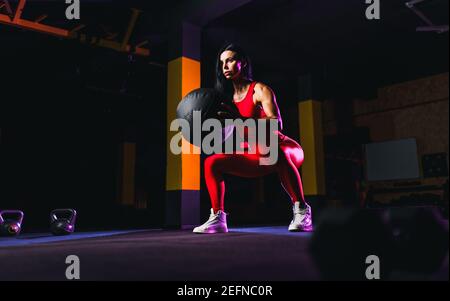 Sportswoman doing squat exercises with fitness ball. Female exercising and stretching with medicine ball at gym. Stock Photo