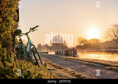 Canal boat with mountain bike left leaning against hedge row early morning sunrise dawn with golden light in sky on the River Trent and mist rising Stock Photo