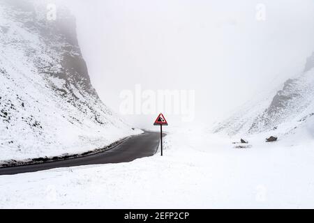 Warning sign red triangle falling rocks on icy snow covered winding mountain road passing through foggy mist filled valley between hills during winter Stock Photo