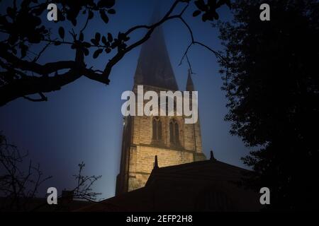 The Chesterfield Crooked Spire of St Marys church lit up illuminated during evening blue hour at night with fog and silhouette of trees Derbyshire Stock Photo