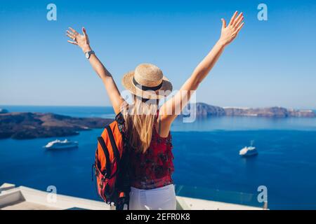 Happy woman traveler walking raising hands in Thera, Santorini island, Greece enjoying sea landscape and cruise ships. Summer vacation Stock Photo