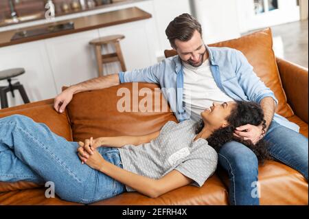 A multiracial couple in love enjoys time together at home, spends a lazy weekend on the couch in cozy living room. African woman lies on the laps of Stock Photo