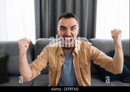 Emotional screaming man watching tv, support and cheering for favorite team. Excited cheerful male sits on a sofa at home, celebrate results of match, or got a great news or deal Stock Photo