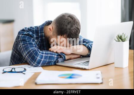 Tired freelancer man is fall asleep at the workplace. Exhausted busy guy working overtime, sits at the desk at home, need a rest, overwork concept. Work from home Stock Photo