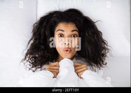 African american young woman lay in bed, posing, making funny faces. Happy female relaxing in bed, after good night sleep. Beautiful girl waking up, feeling fresh and cheerful at the morning Stock Photo