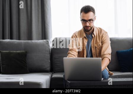 Concentrated male freelancer working from home, searching information in the network. Caucasian male manager or IT specialist sitting on the couch, using laptop, chatting online, distant work Stock Photo