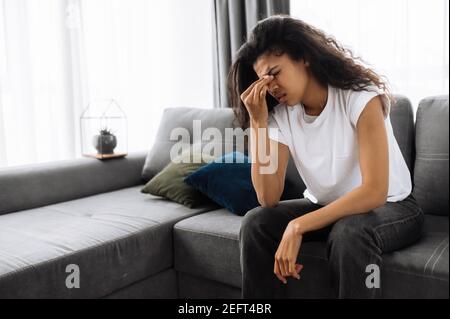 Tired girl sitting on the sofa at home, having a terrible headache. Exhausted african american female massage the eyes, feeling unhealthy. Young woman catch a cold or having flu, upset about illness Stock Photo