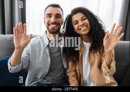 Multiracial couple in love, caucasian guy and african american girl, sitting on a sofa talk on video call with friends or family greeting, smiling and waving hands Stock Photo