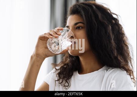 Drink water every day. African american young woman drinks a glass of pure fresh water while sitting on the sofa at home. Beautiful healthy woman follow healthy lifestyle Stock Photo