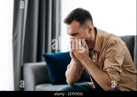 Unhealthy young man. Caucasian guy is sick with flu, he sneezes and blows his nose in a napkin while sitting on the couch at home Stock Photo