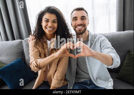 Multiracial couple in love sits on the couch at home and shows a heart gesture with their hands. Love and relationship concept Stock Photo