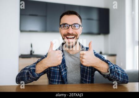 Portrait of a friendly caucasian student or freelancer guy during video call, he looks at the camera, smiling and and gesturing with hands showing thumbs up Stock Photo
