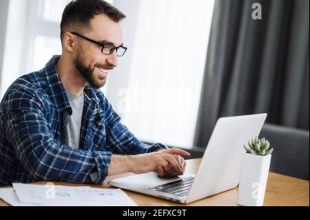 Smiling freelancer guy works from home, using laptop, chatting with colleagues online. Handsome caucasian busy man is developing new project, searching ideas or plans for startup Stock Photo