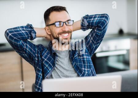 Positive freelancer guy relaxing at the workplace and smiling. Confident caucasian male entrepreneur sitting at home office, taking break from distant working and dreaming about vacation Stock Photo