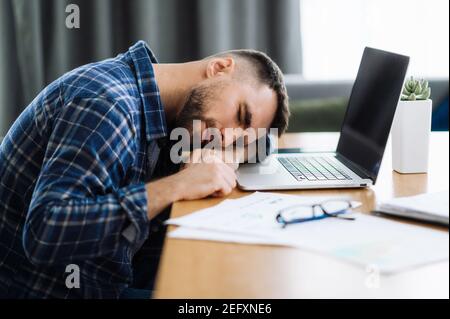 Tired freelancer man is fall asleep at the workplace. Exhausted busy guy working overtime, sits at the desk at home, need a rest, overwork concept. Work from home Stock Photo